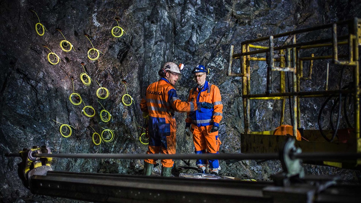 To mannlige fagarbeidere stående inne ved en fjellvegg i en tunnel